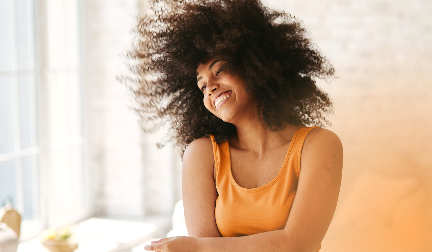 Lachende Frau mit Afrohaar in orangefarbener Kleidung steht locker in einem hellen Wohnzimmer.