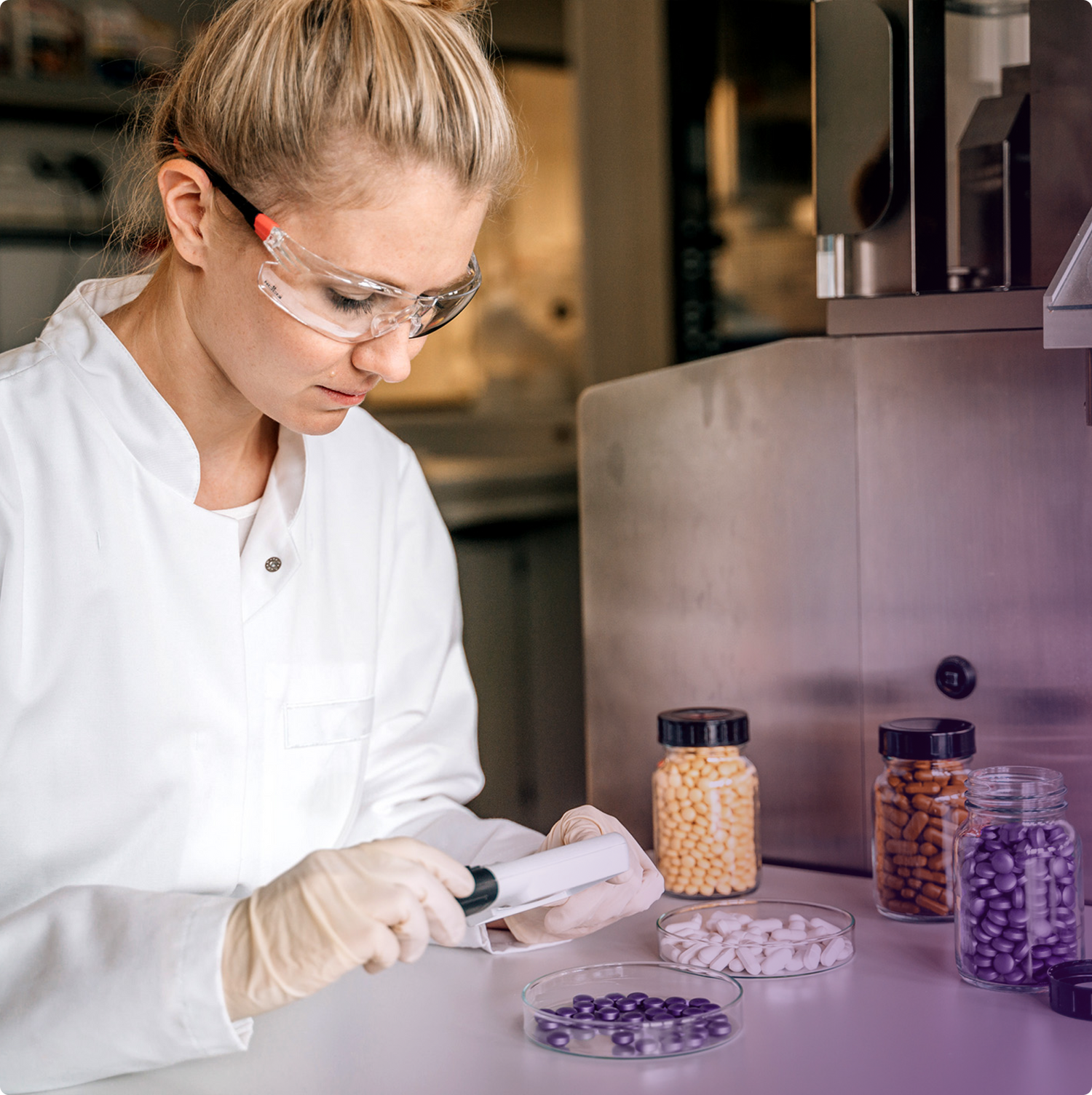 Eine Frau in einem weißen Laborkittel und mit Schutzbrille arbeitet in einem Labor. Sie trägt Handschuhe und untersucht mit einem Handscanner Tabletten in einer Petrischale. Auf dem Tisch stehen mehrere Gläser mit verschiedenfarbigen Kapseln und Tabletten.