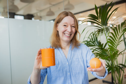 Eine Frau mit schulterlangen, rötlich-blonden Haaren steht in einem modernen Büro mit Pflanzen im Hintergrund. Sie trägt ein hellblaues Oberteil und lächelt freundlich. In der linken Hand hält sie eine orangefarbene Produktverpackung in Zylinderform, in der rechten eine dazu passende orangefarbene Kugel mit der Aufschrift „rund.um“.