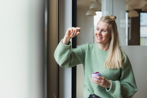 Eine Frau mit langen, blonden Haaren trägt einen grünen Strickpullover und steht an einem Fenster in einem modernen Büro. Sie lächelt und hält in der linken Hand einen geöffneten, lilafarbenen Behälter mit kleinen, roten Kapseln. Mit der rechten Hand hält sie eine einzelne Kapsel hoch.