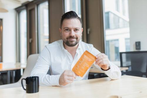 Ein Mann mit dunklen, zurückgekämmten Haaren und Bart sitzt in einem modernen Büro an einem Tisch. Er trägt ein weißes Hemd und lächelt leicht in die Kamera. In den Händen hält er eine orangefarbene Verpackung mit der Aufschrift „rund.um energy“. Auf dem Tisch steht eine schwarze Tasse.