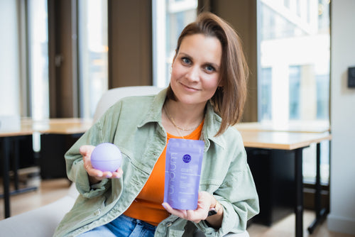 Eine Frau mit halblangen, braunen Haaren sitzt in einem modernen Büro und lächelt in die Kamera. Sie trägt ein orangefarbenes Oberteil unter einer hellgrünen Jacke. In der linken Hand hält sie eine lilafarbene Kugel, in der rechten ein lilafarbenes Produkt mit der Aufschrift „calm base“ der Marke „rund.um