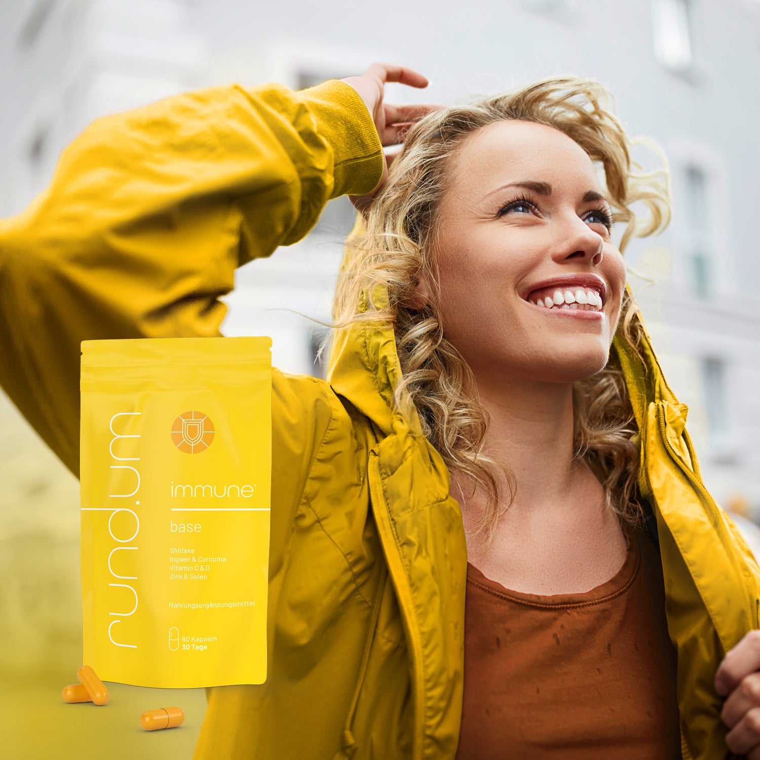 Eine junge Frau mit lockigem Haar trägt eine gelbe Jacke mit Kapuze und lacht strahlend, während sie durch eine Stadtstraße geht. Im Hintergrund sind unscharf Häuserfassaden und Autos zu sehen.