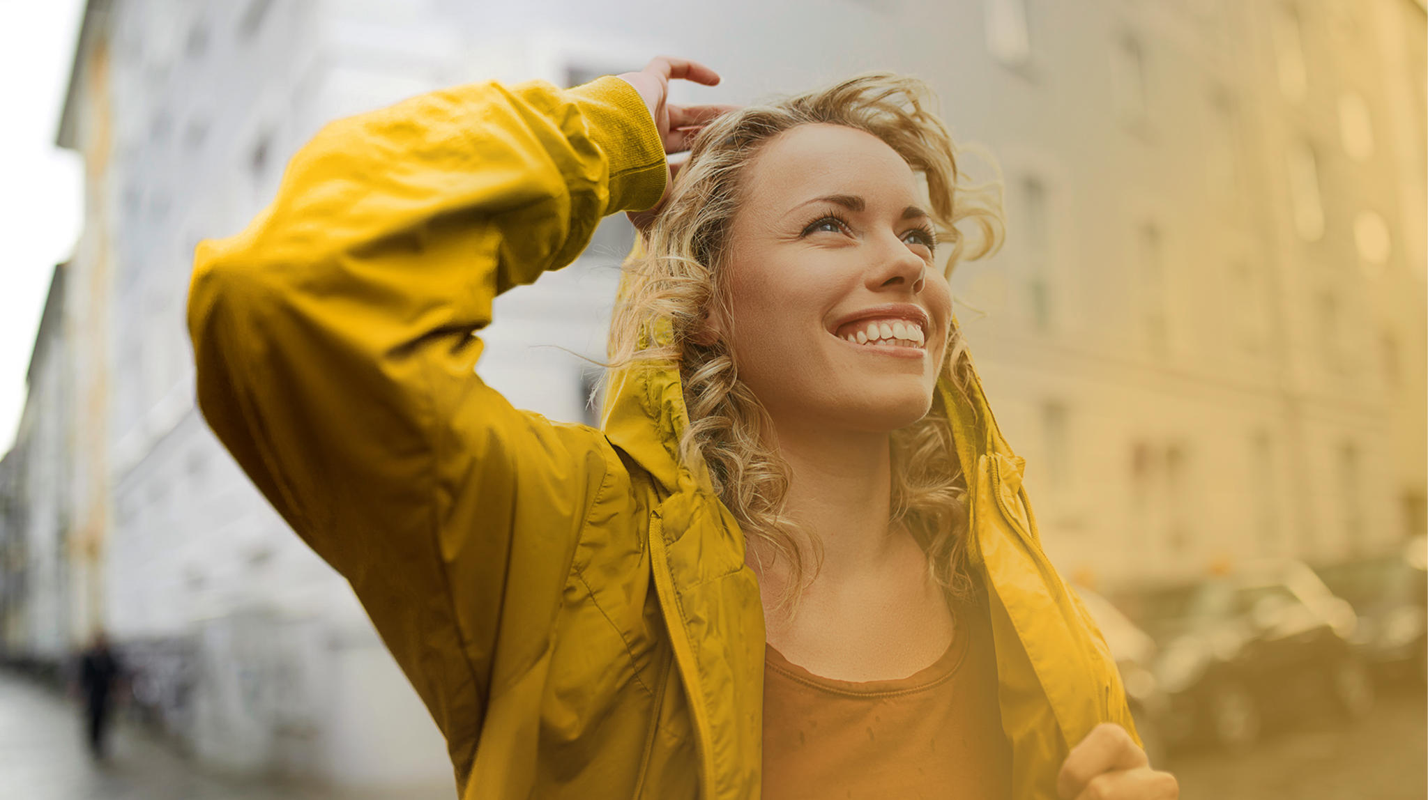 Eine junge Frau mit lockigem Haar trägt eine gelbe Jacke mit Kapuze und lacht strahlend, während sie durch eine Stadtstraße geht. Im Hintergrund sind unscharf Häuserfassaden und Autos zu sehen.