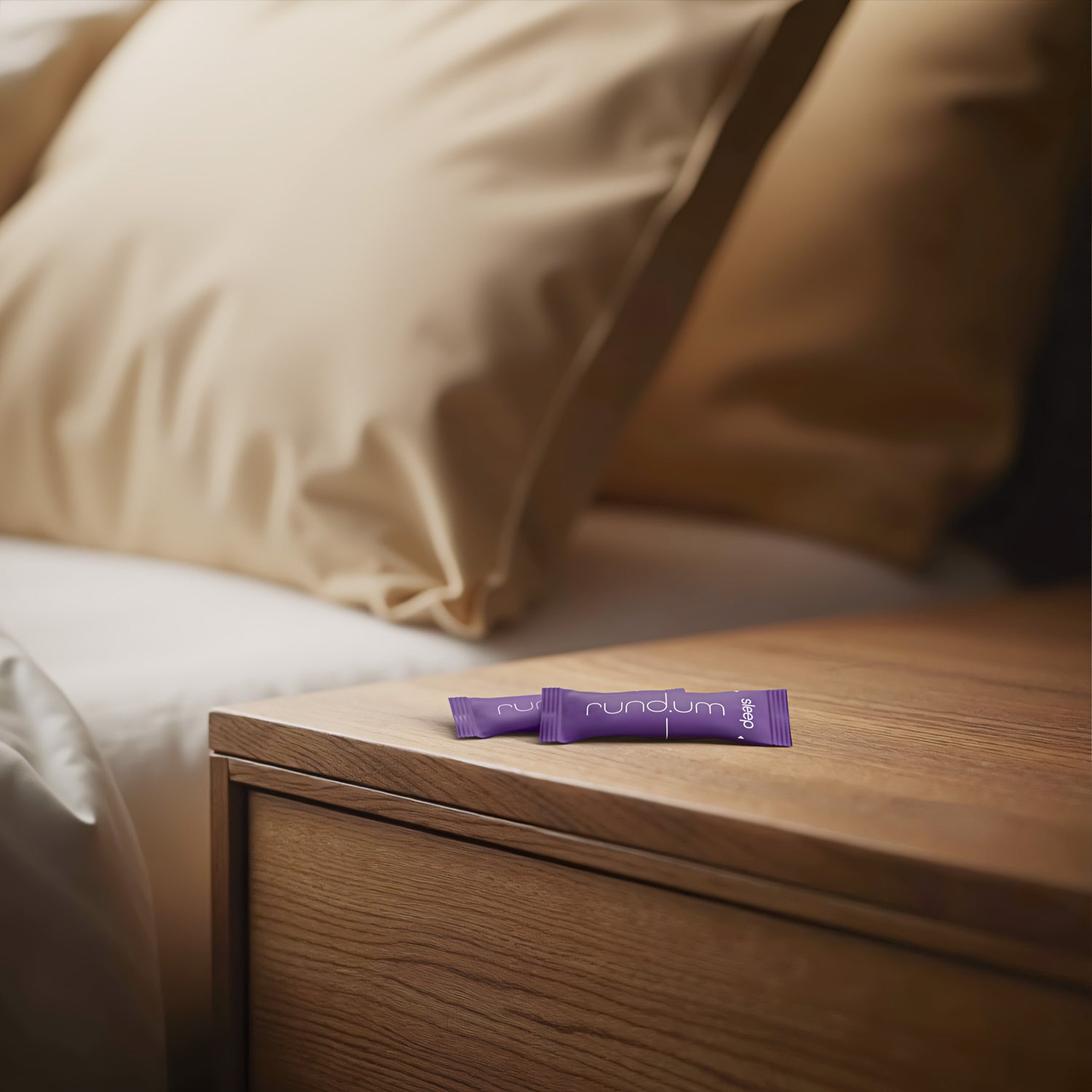 Zwei Stick-Packungen ‚sleep plus‘ von rundum auf einem Nachttisch neben einem Bett mit beigefarbenen Kissen